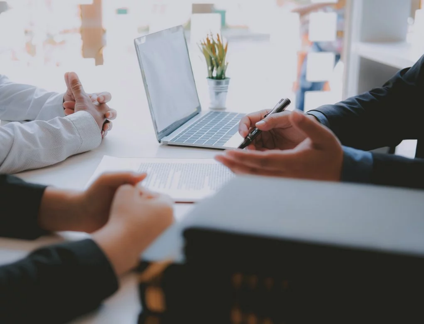 Szene eines Business-Meetings. Die Arme von drei Personen sind sichtbar. Eine Person hält einen Stift. Im Vordergrund ein Stapel Bücher, im Hintergrund ein aufgeklappter Laptop.